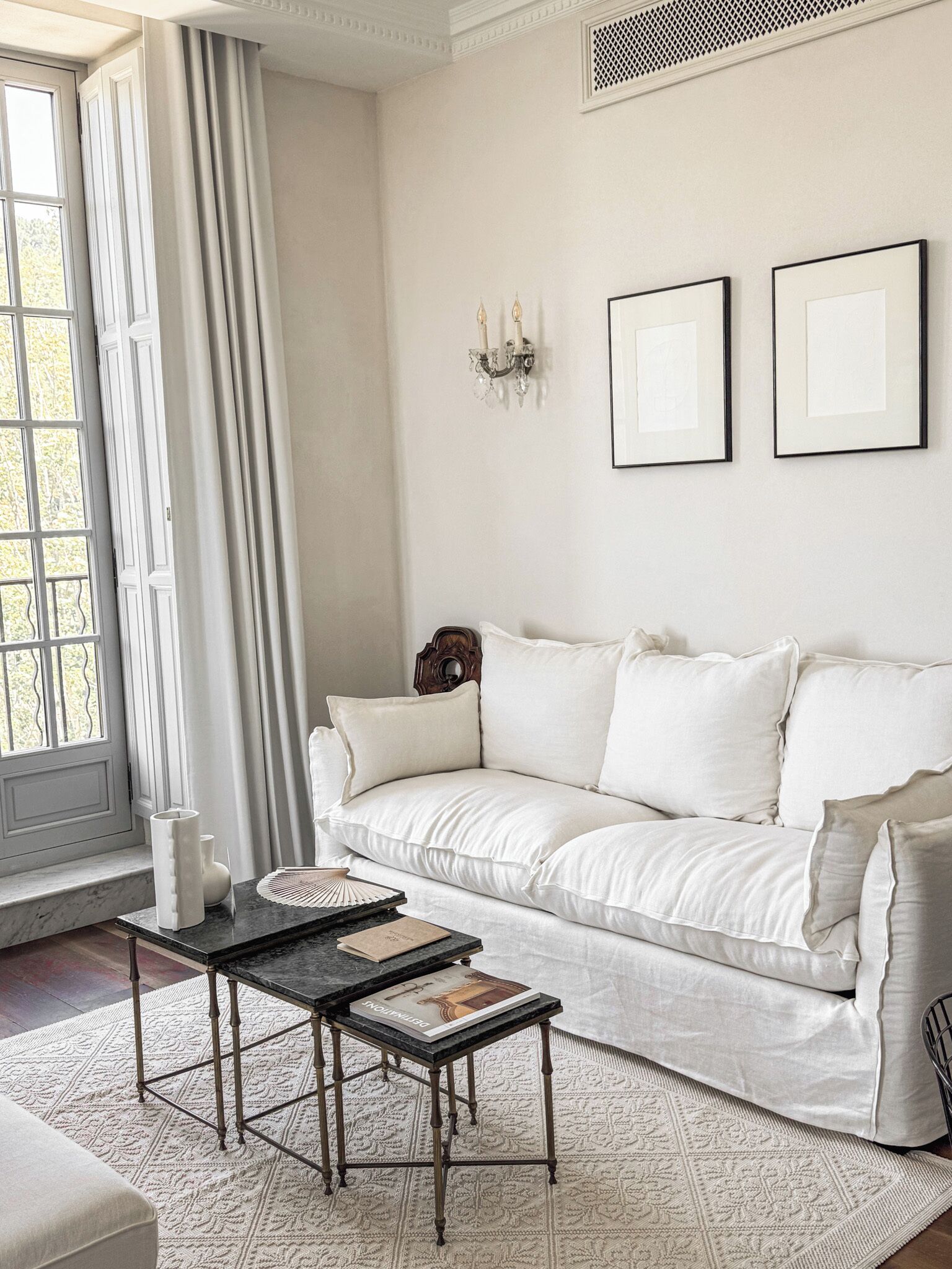 Sala de estar com um sofá branco, uma mesa de centro preta e um tapete estampado. Há uma janela com cortinas, um aplique de parede e duas molduras com quadros.