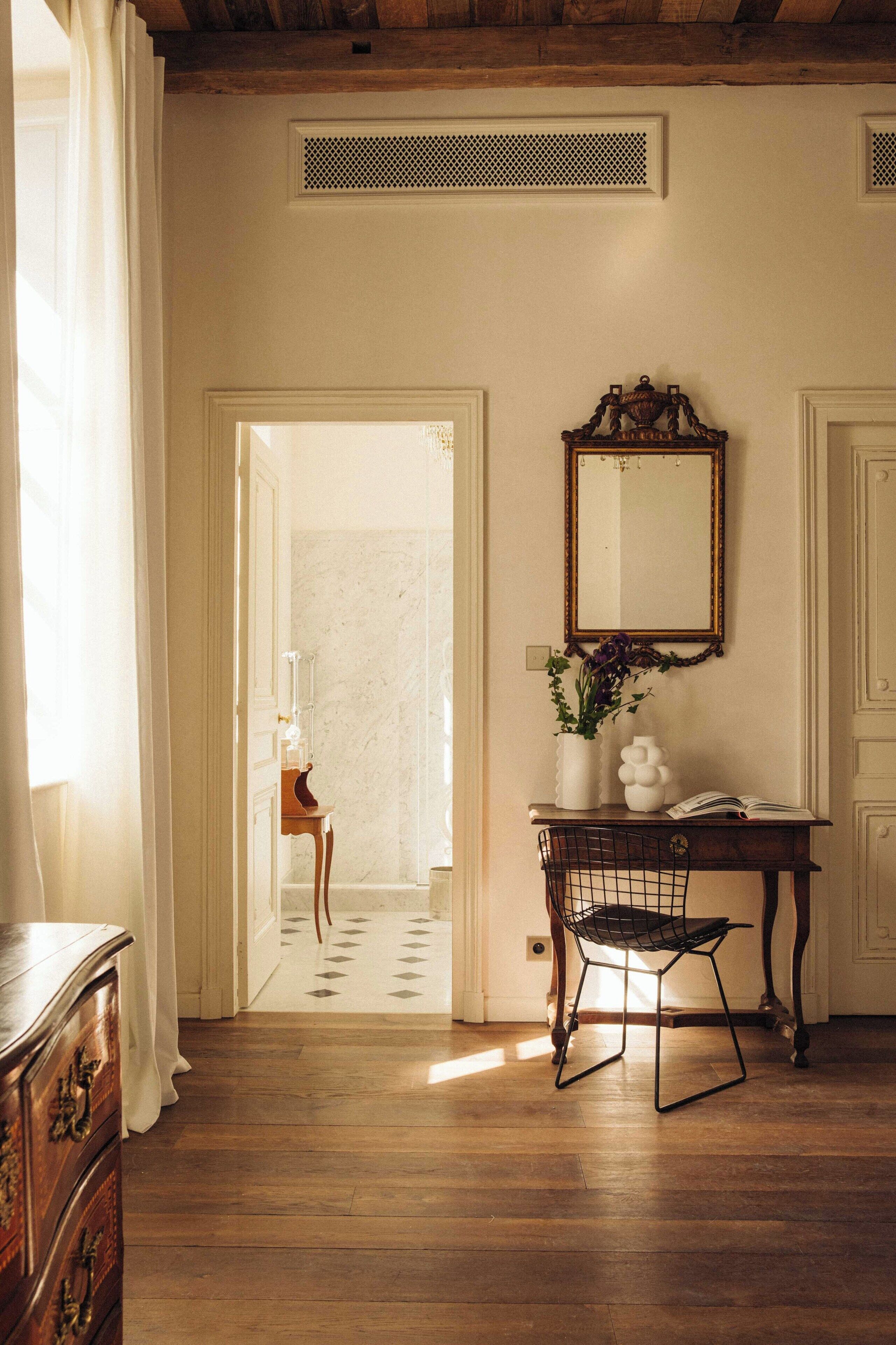 Una habitación con piso de madera, un escritorio con silla, un espejo y un florero con flores.