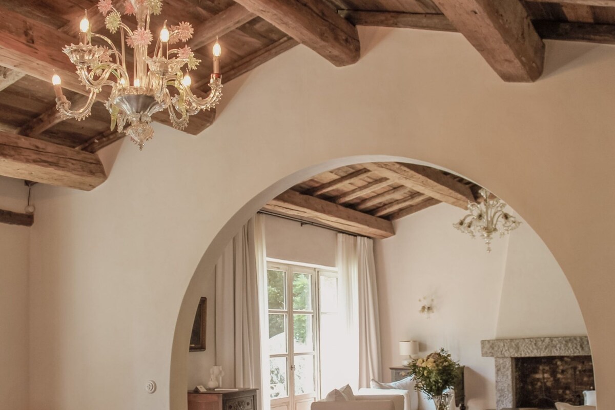 A spacious living room with a high wooden ceiling, a chandelier, a fireplace, and a large arched doorway leading to another room.