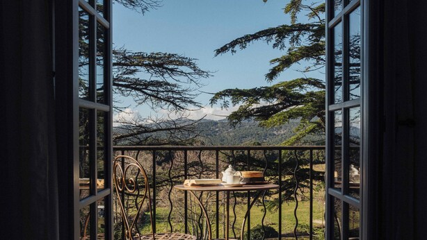 Un balcon ouvert, agrémenté d’une chaise et d’une table en fer forgé, d’où l’on peut admirer un paysage montagneux.