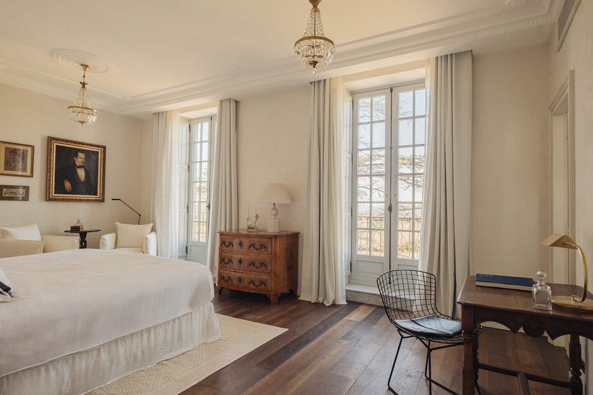 Een ruime slaapkamer met een groot bed, een houten dressoir, een bureau met stoel en een kroonluchter.