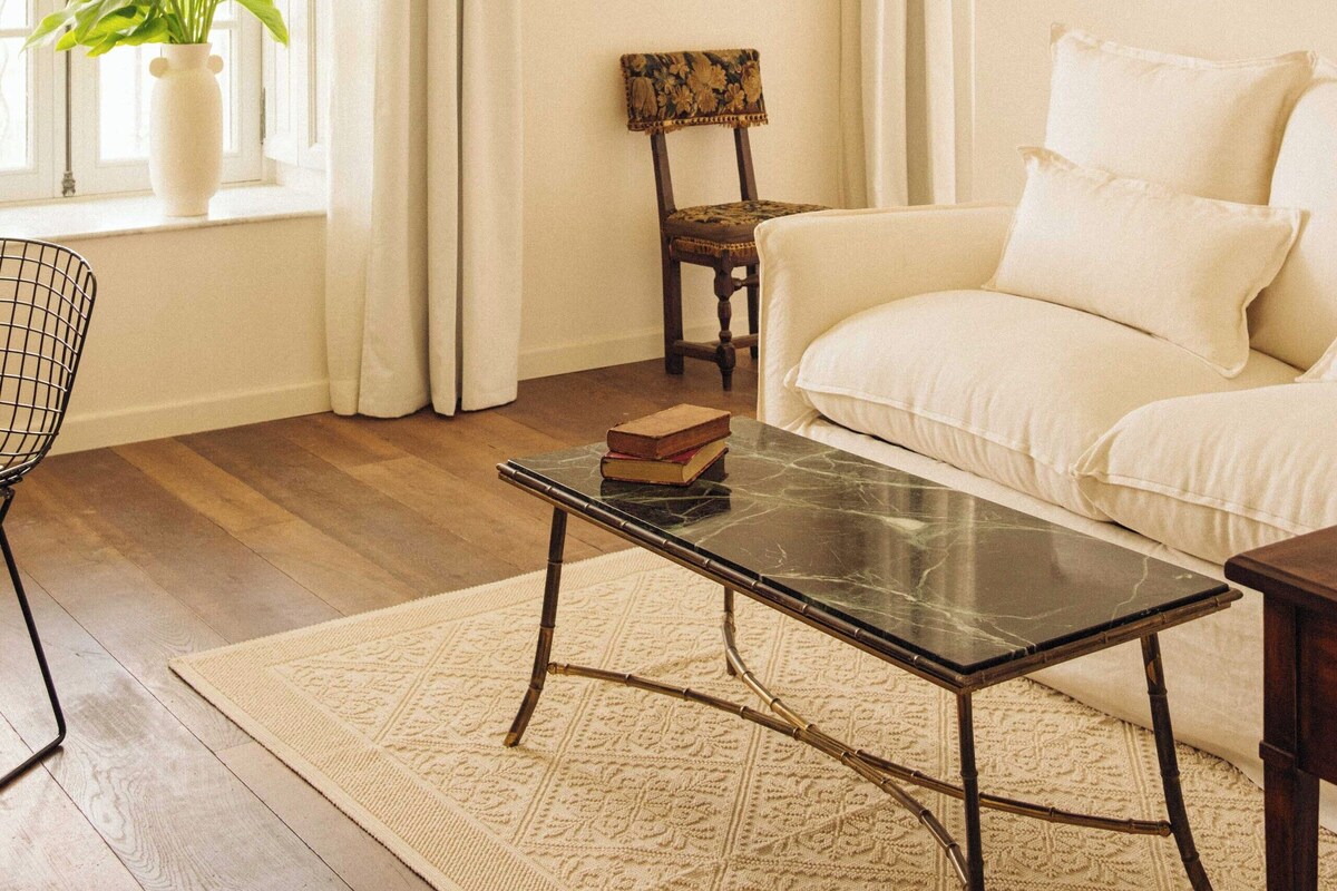 A room with a white sofa, a black coffee table, a white vase with flowers, a wooden chair, and a chandelier.
