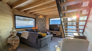 A cozy cabin interior with a stone wall, wooden beams, a sofa, a TV, and a staircase. - Nature Chalet with Sauna and WIFI near Gérardmer (Anould)