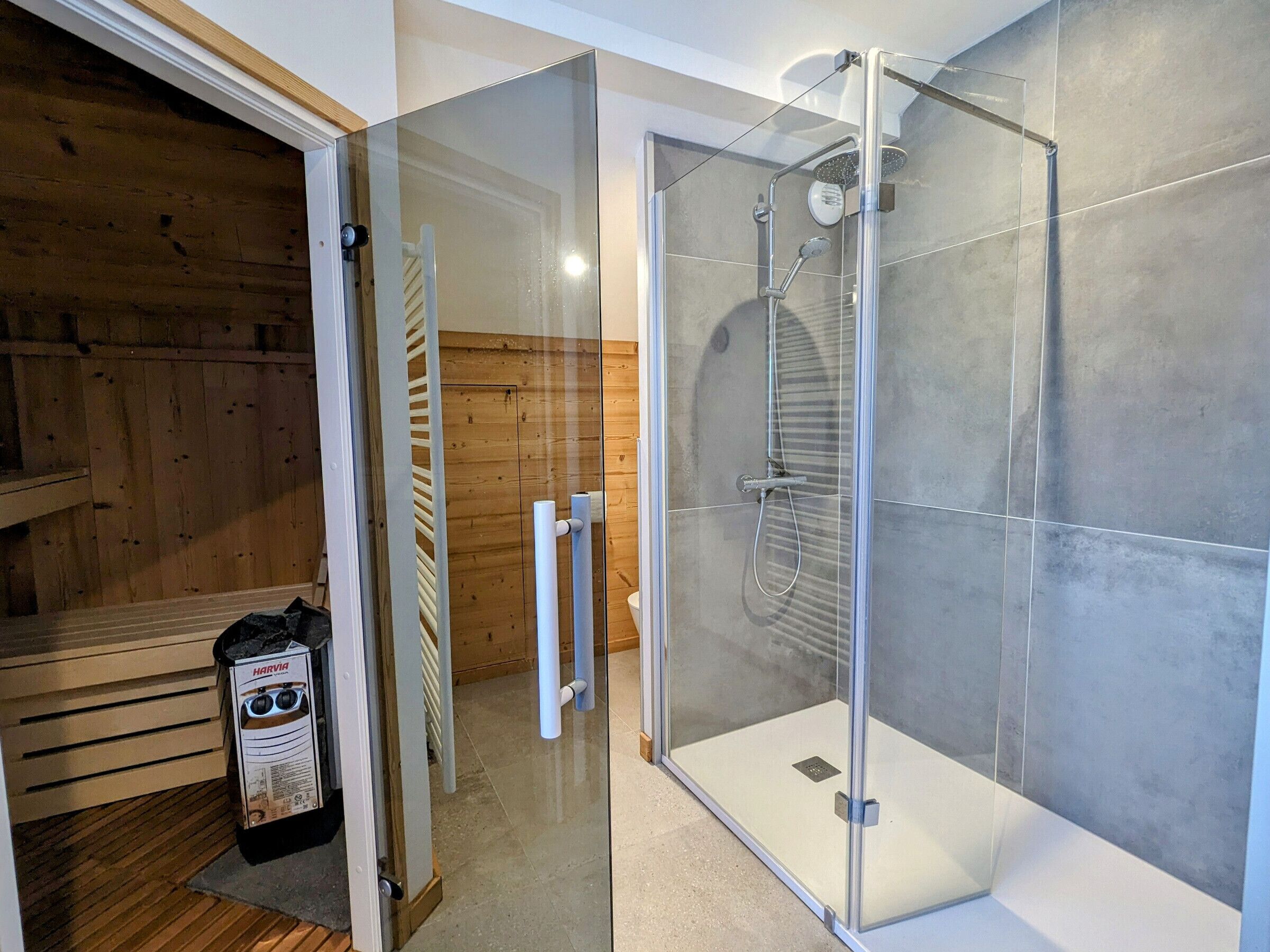 Un baño moderno con ducha envuelta en vidrio, una sauna de madera y un calentador de sauna Harvia.