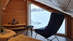 A cozy cabin interior with a large window offering a snowy landscape view, a comfortable chair, and a small table with a plant. - Nature Chalet with Sauna and WIFI near Gérardmer (Anould)