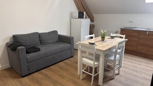 A compact living space with a grey sofa, a wooden dining table with white chairs, a small kitchen area with a microwave, and a radiator. - Country Escape: Renovated Cottage with Pets Allowed (Saint-Bonnet-de-Four)