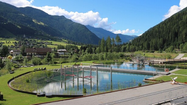 A swimming pool with a bridge, surrounded by a mountainous landscape and a small village.