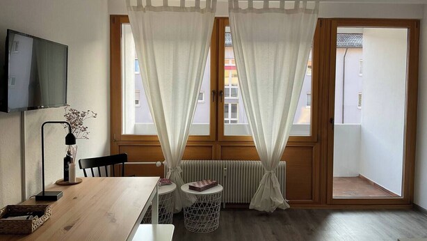 A modern room with a wooden dining table, two small round tables, a radiator, and a large window with sheer curtains. - Renovated loft with parking space and balcony (Lörrach)