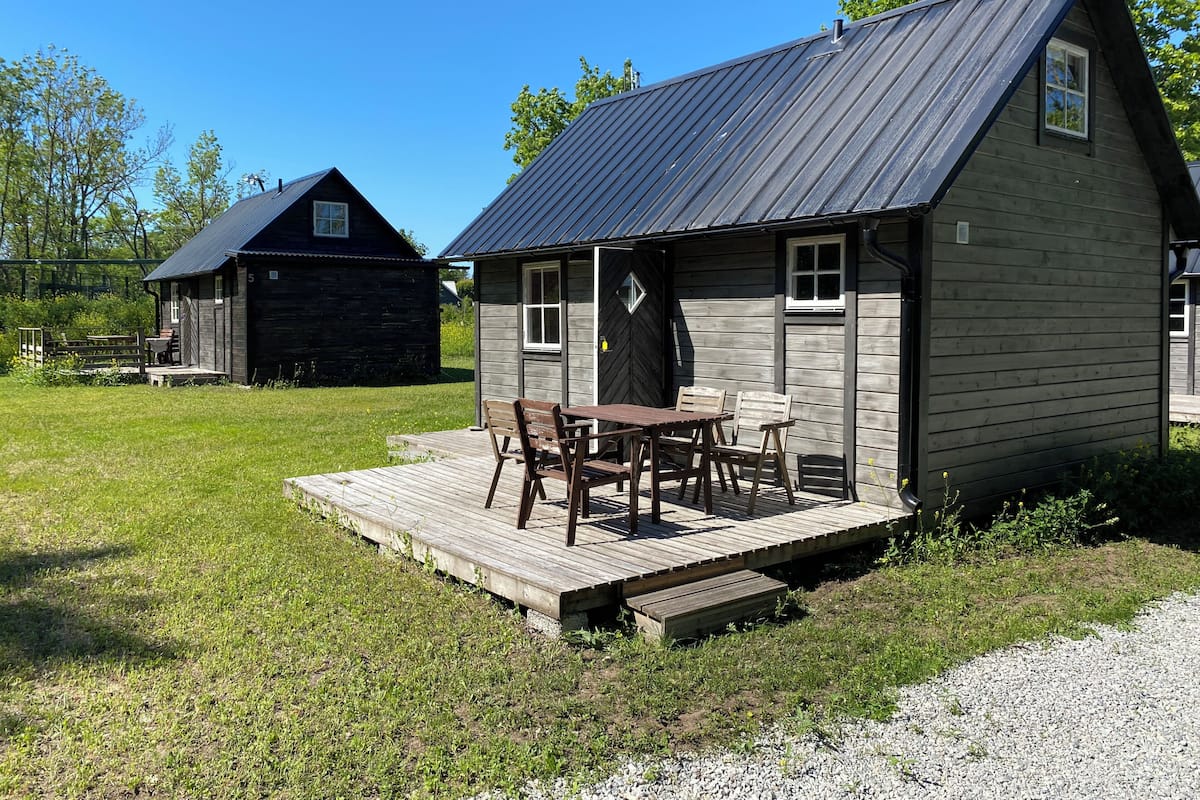 A wooden cabin with a porch and a table, surrounded by a grassy area and another building in the background.