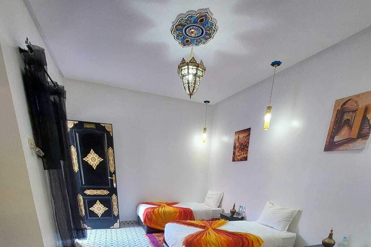 Et hotelværelse med to senge, et rødt tæppe, et gulv med mønster, en dekorativ loftlampe og en sort dør med guld detaljer.