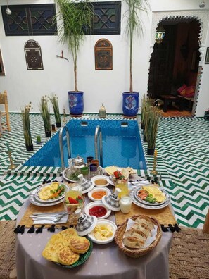 A dining table set with various dishes, a pool, and decorative elements in a room with patterned flooring and traditional decor. - Riad Zaid (Marrakech)