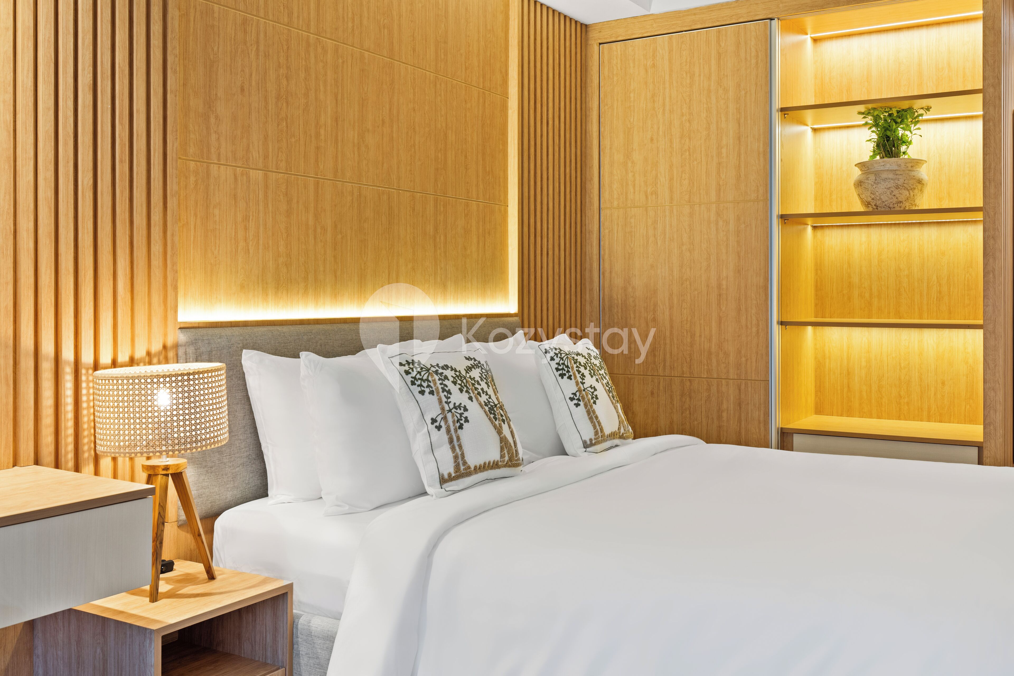 Ein modernes Hotelzimmer mit einer Wandpaneele aus Holz, einem Nachttisch mit Lampe, einem Bett mit weißer Bettwäsche und Dekokissen sowie einem Regal mit einer Pflanze.