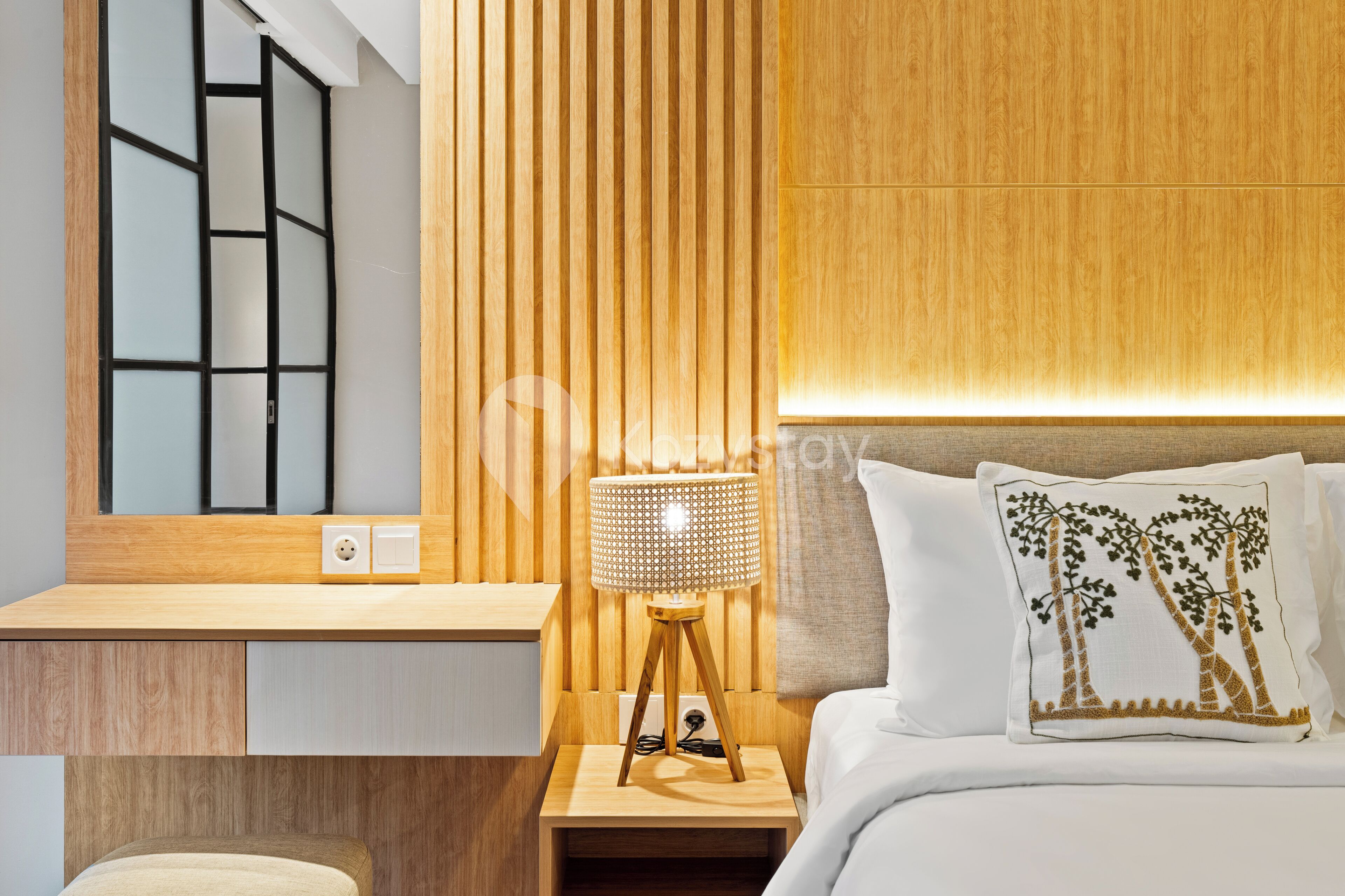 A modern hotel room with a wooden headboard, a bedside lamp, a nightstand, and a decorative pillow.