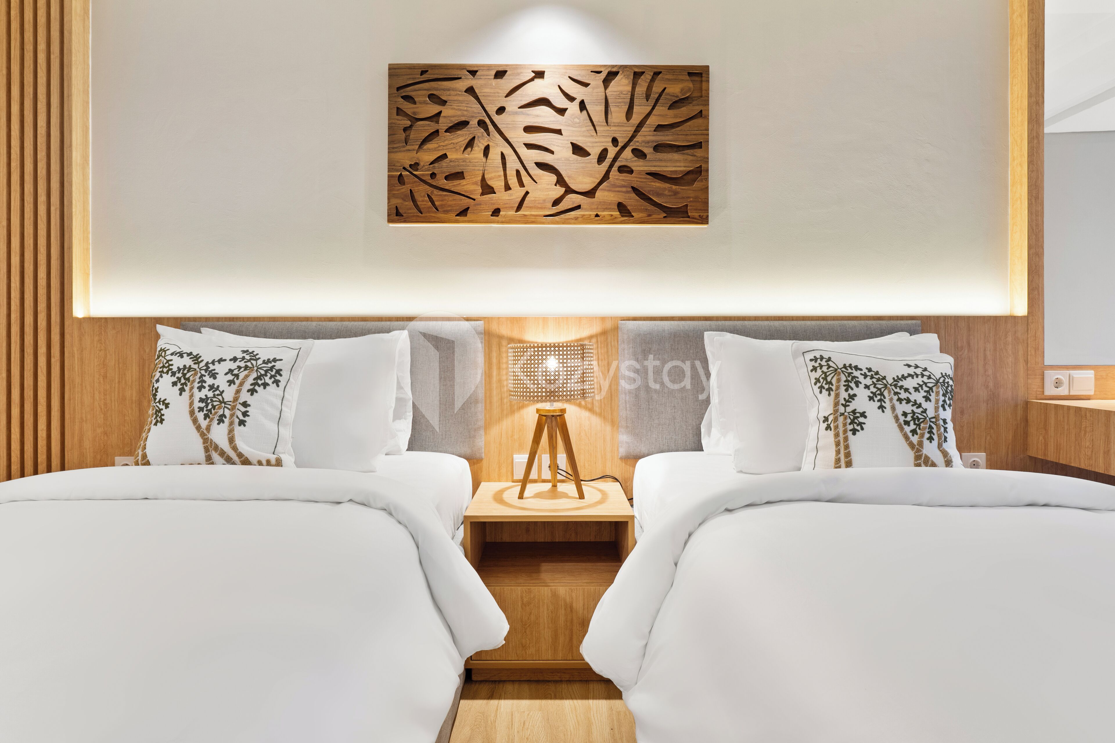 Quarto de hotel com duas camas, cabeceira de madeira, uma obra de arte na parede e uma mesa pequena com abajur.