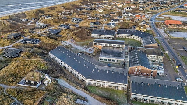 Ovenfra set en kystby med en blanding af boliger og moderne bygninger, heriblandt en stor, hvid bygning med fladt tag og mange vinduer.