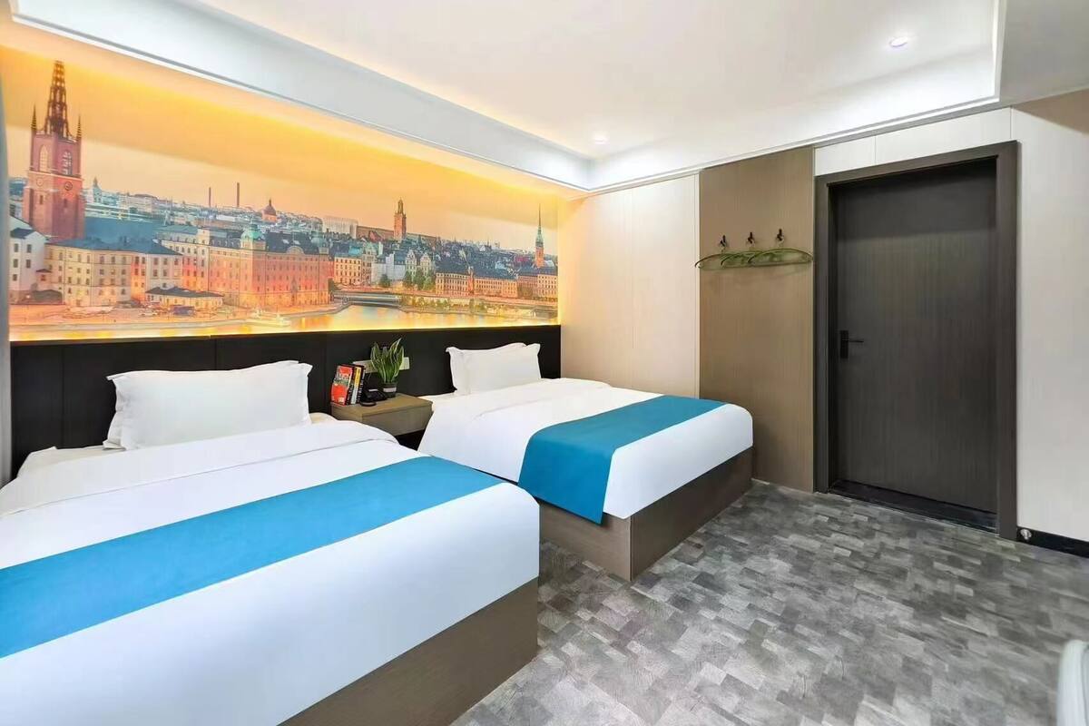 A hotel room with two beds, a cityscape mural, a wooden door, and a small table with a plant.