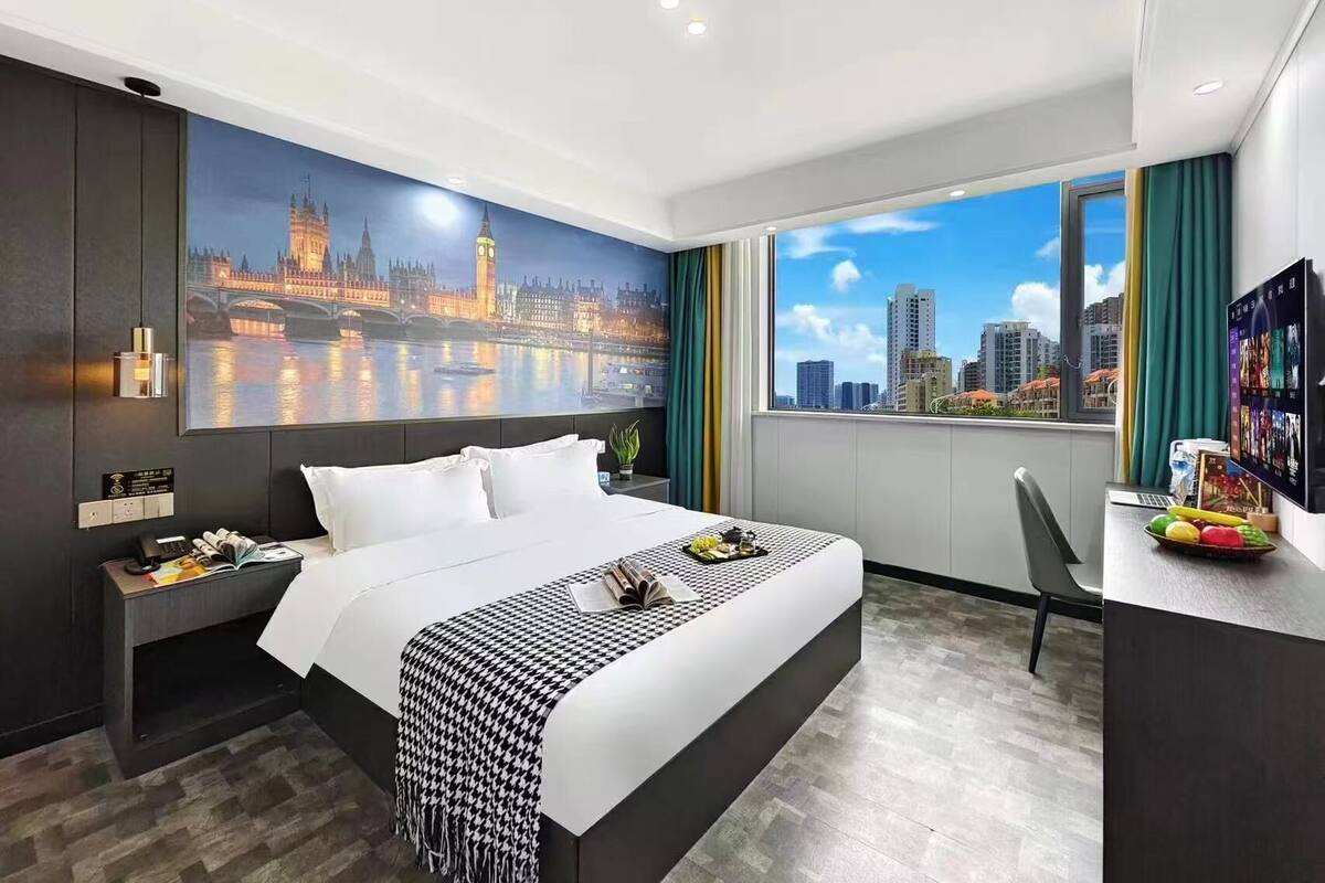 A hotel room with a large bed, a desk with a laptop, a view of a cityscape, and a painting of a famous bridge.