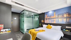 Una habitación de hotel moderna con cama, escritorio con computadora portátil, ducha con mampara de vidrio y un mural con vista a la ciudad en la pared.