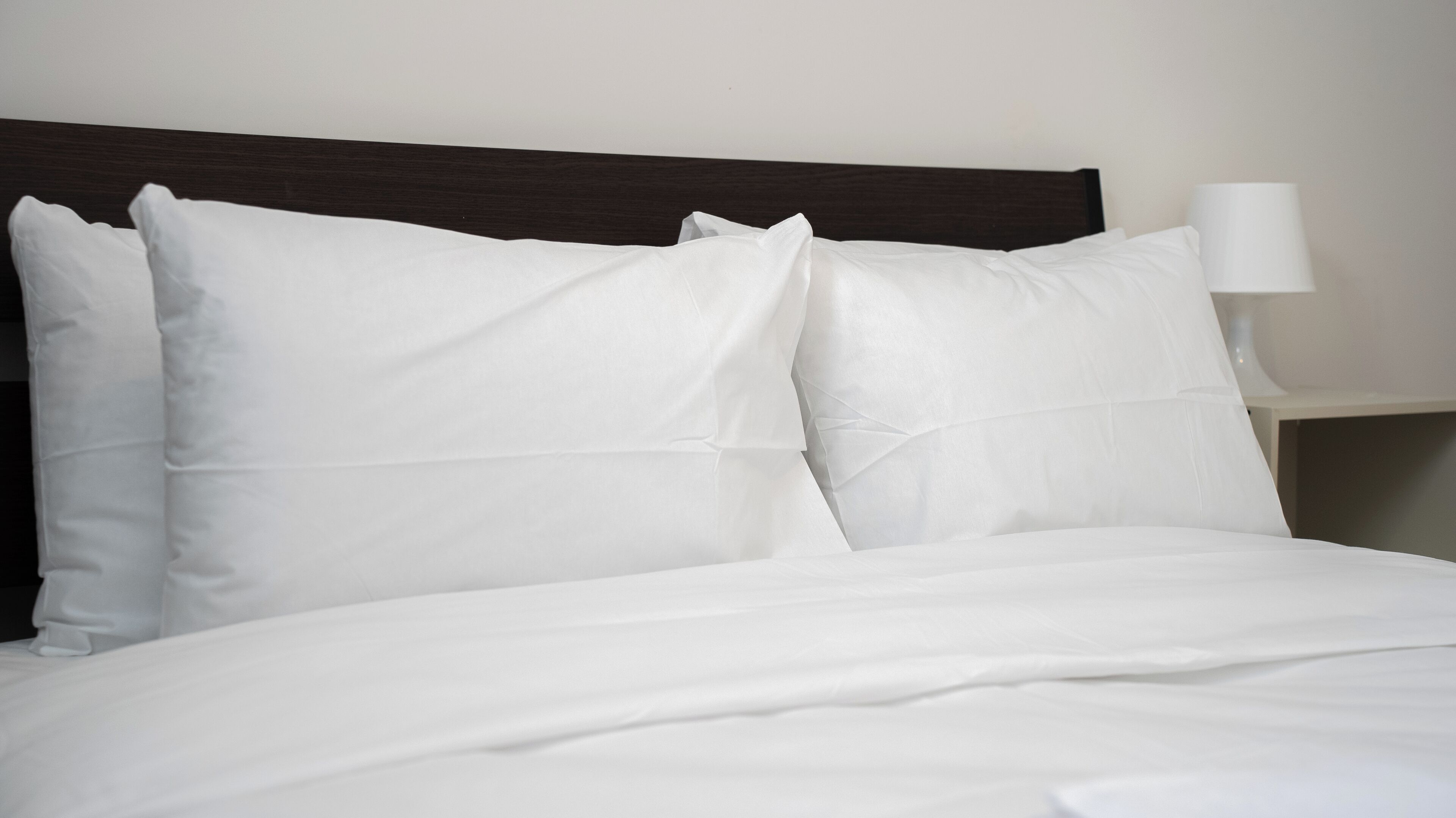 Un lit bien fait, avec du linge de lit blanc et des oreillers, une tête de lit en bois foncé et une lampe de chevet posée sur une table de chevet blanche.