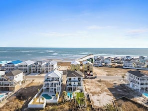 A coastal residential area with multiple two-story houses, a beach, and a swimming pool. - The Grand View by Brunswick Vacation Rentals: New 5BR Home w/ Elevator & Pool (Ocean Isle Beach)