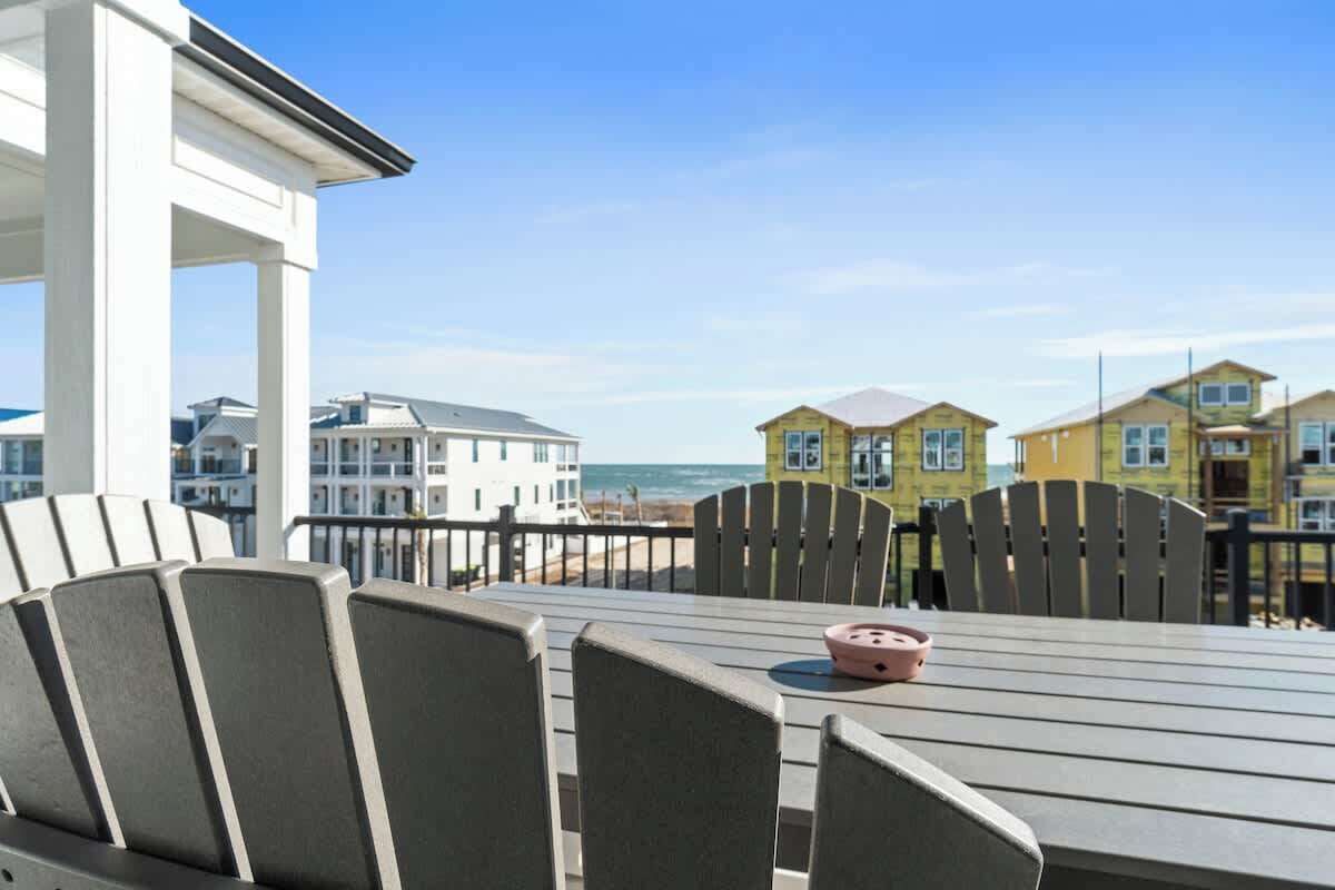 Een terras met een tafel en stoelen, met uitzicht op een strand met gele huizen en de oceaan.