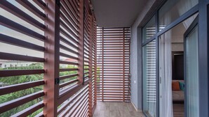 Un balcone con vista su vegetazione e edifici, una parete divisoria in legno a lamelle e una porta in vetro che dà su una stanza con un letto e cuscini.