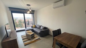 A modern living room with a sectional sofa, a wooden dining table, and a view through large windows. - I&I Apartments III (Kragujevac)