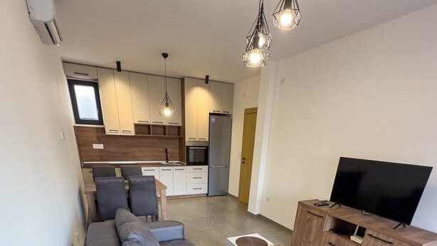A modern living room with a grey sectional sofa, a wooden TV stand, and a kitchen in the background. - I&I Apartments III (Kragujevac)