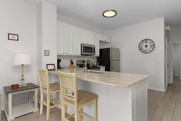 A modern kitchen with white cabinets, a central island with bar stools, stainless steel appliances, and a wall clock.