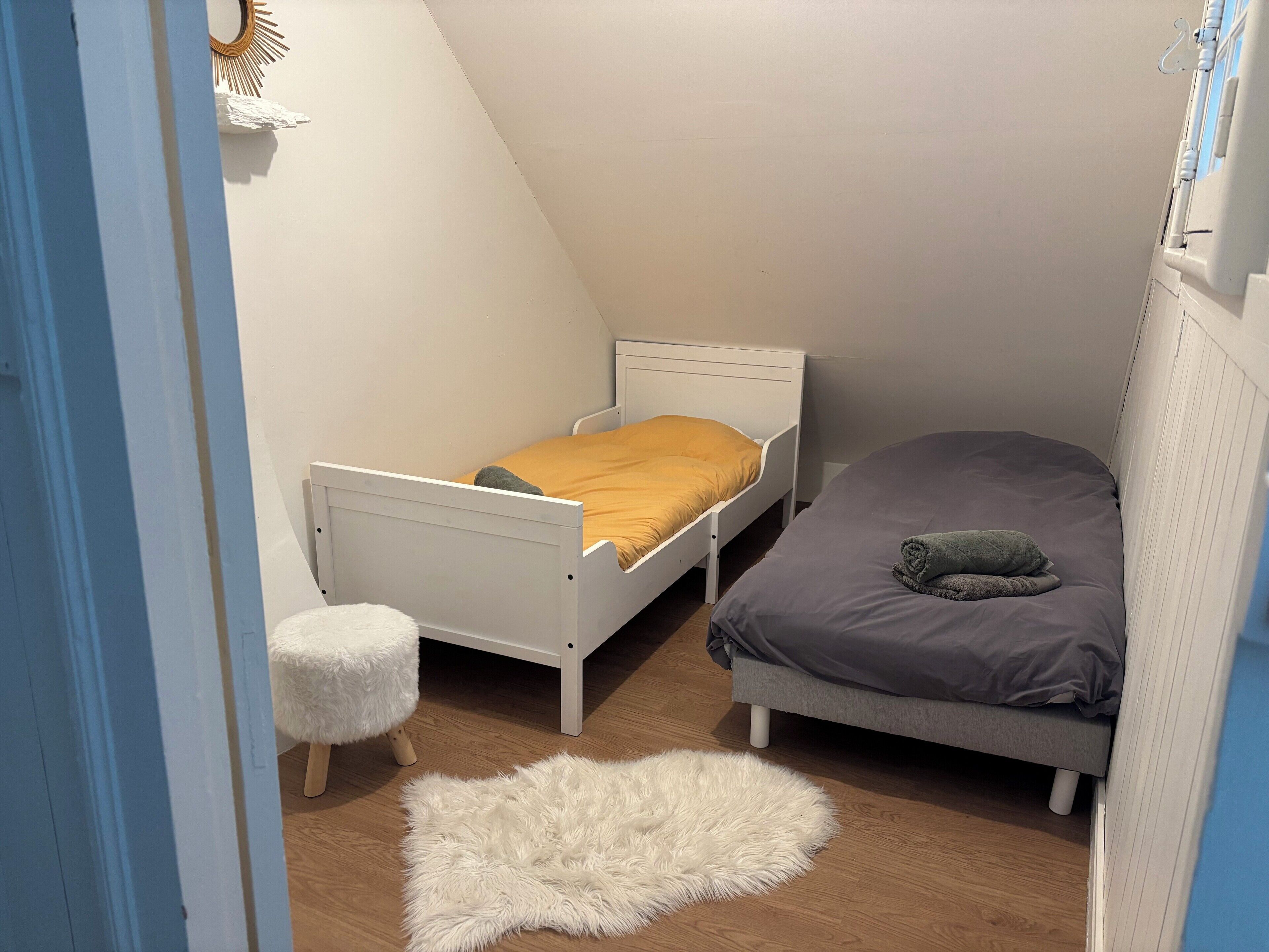 A small, triangular room with a white bed, a gray bed, and a white furry rug on the floor.