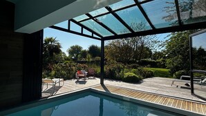 Piscina interior moderna con techo de cristal, rodeada por una terraza de madera y asientos exteriores.