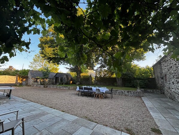 An outdoor dining area with a table set for four, surrounded by stone walls and a large tree providing shade.