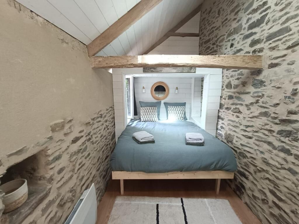 Een slaapkamer met een stenen muur, een houten balk in het plafond, een bed met een blauw beddegoed en een ronde spiegel boven het bed.