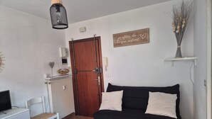 A compact living space with a black sofa, a white chair, a small table, a radiator, a wooden door, and a wall-mounted sign. - Studio per week  (Menton)
