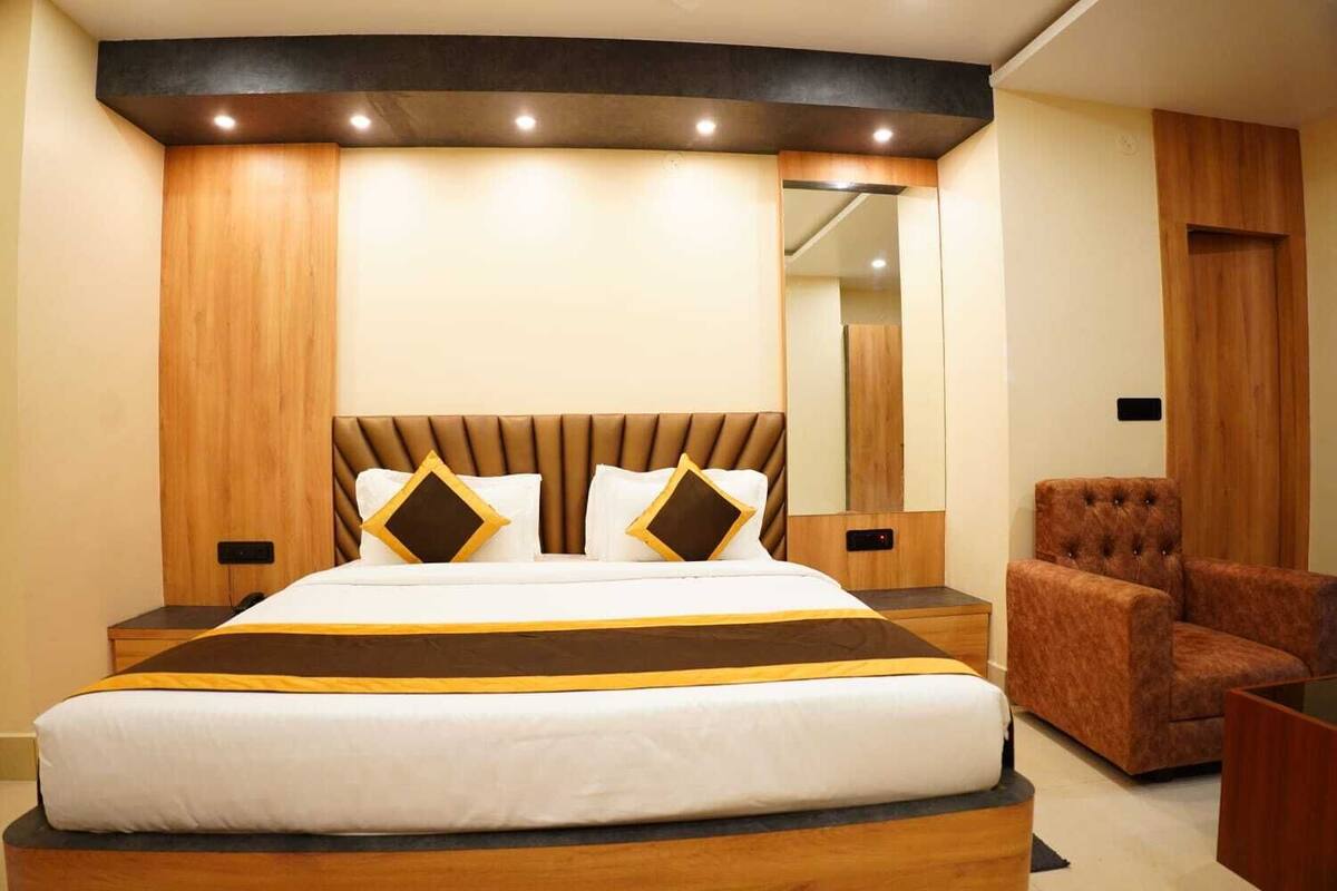 A modern hotel room with a large bed, a wooden headboard, a brown upholstered armchair, and a nightstand with a lamp.