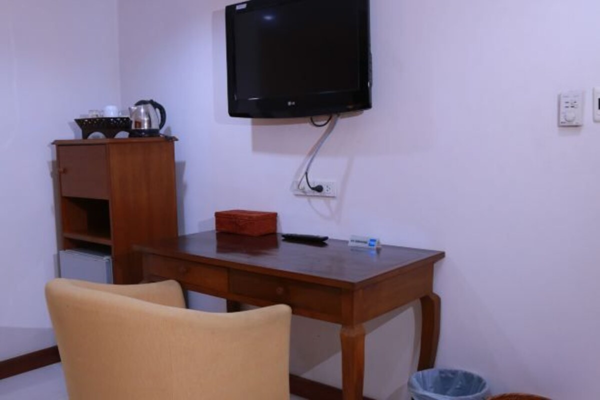 Una habitación pequeña con un escritorio de madera, un televisor montado en la pared, una silla, un cesto de basura y un mueble pequeño con una tetera encima.