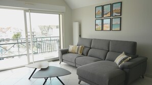 Een woonkamer met een grijze bank, een bijzettafeltje en een balkon met uitzicht op gebouwen.