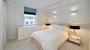 Une chambre moderne avec un grand lit, une commode blanche, des lampes de chevet et une fenêtre donnant sur des immeubles.