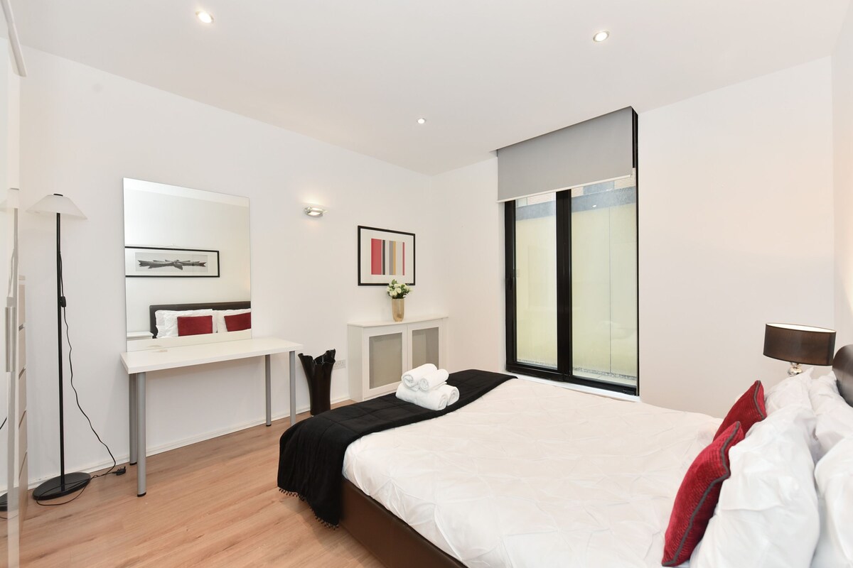 A modern hotel room with a large bed, a desk, a chair, a floor lamp, and a window with blinds.