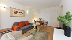 A modern living room with a grey sofa, orange cushions, a glass coffee table, and a dining area with chairs and a table. - Cromwell Style I - 605pw (London)