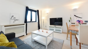 Una sala de estar moderna con un sofá blanco, una mesa de centro blanca, un televisor y un comedor con una mesa y sillas de madera.