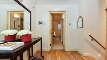 A hallway with a wooden floor, a dark wooden console table with a magazine and red flower pots, a bench, and a mirror on the wall.