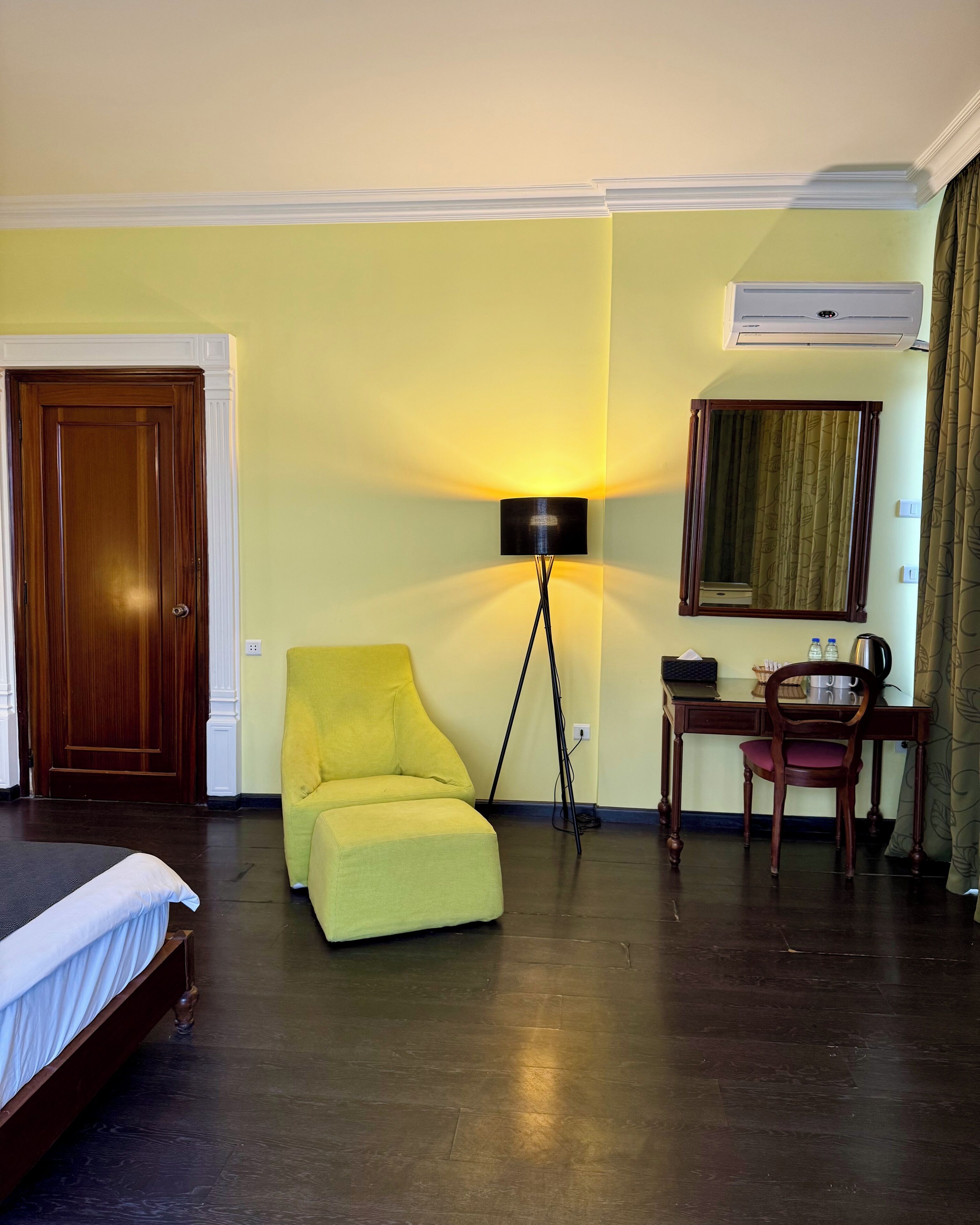 Una habitación de hotel con una cama, un sillón amarillo, un escritorio con una silla, una lámpara de pie, un equipo de aire acondicionado y un espejo.