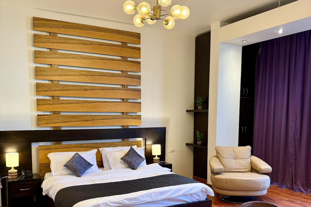 A modern bedroom with a large bed, wooden headboard, a chandelier, a leather armchair, and a desk with a chair.