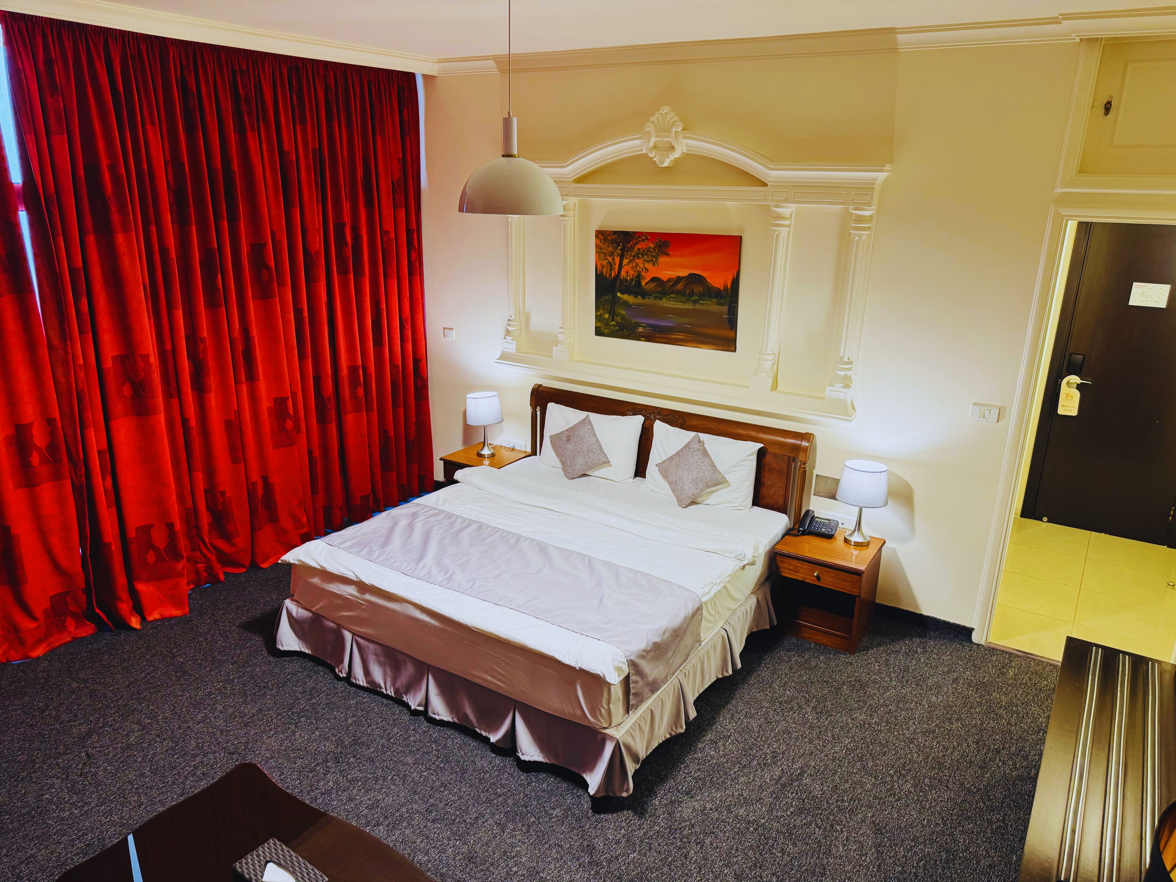 A hotel room with a large bed, two bedside tables, a painting above the bed, and a door to the right.