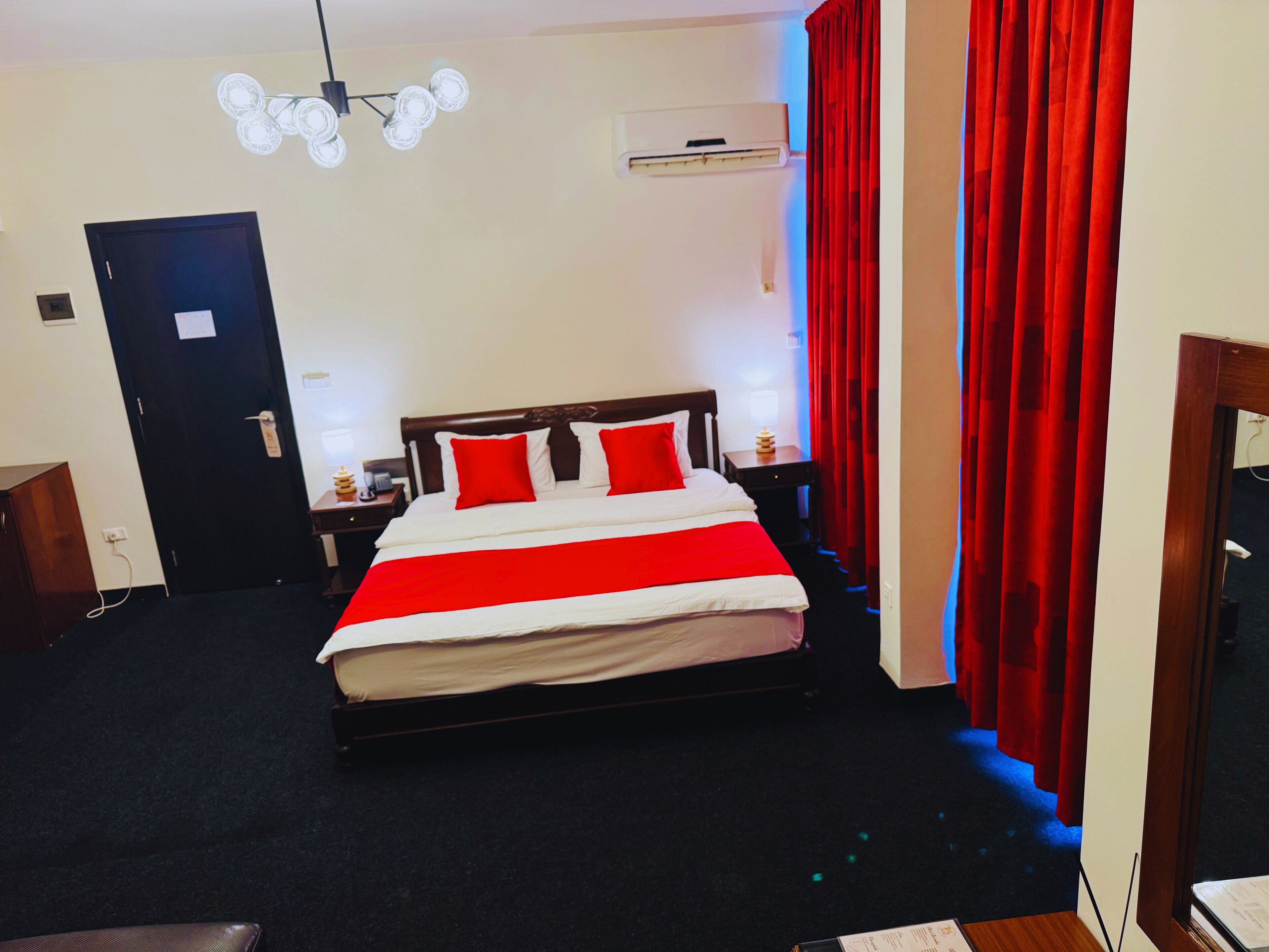 A hotel room with a bed, a nightstand, a lamp, a ceiling light fixture, and a door.