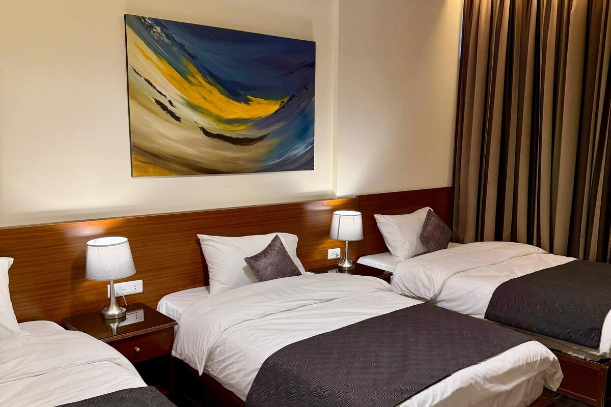 A hotel room with three beds, a wooden headboard, a painting on the wall, and a curtain on the window.