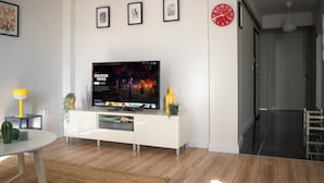 A modern living room with a flat-screen TV displaying 'Stranger Things', a white entertainment unit, a yellow lamp, and a red wall clock. - Cozy flat in Kadıköy 2BR New House (İstanbul)