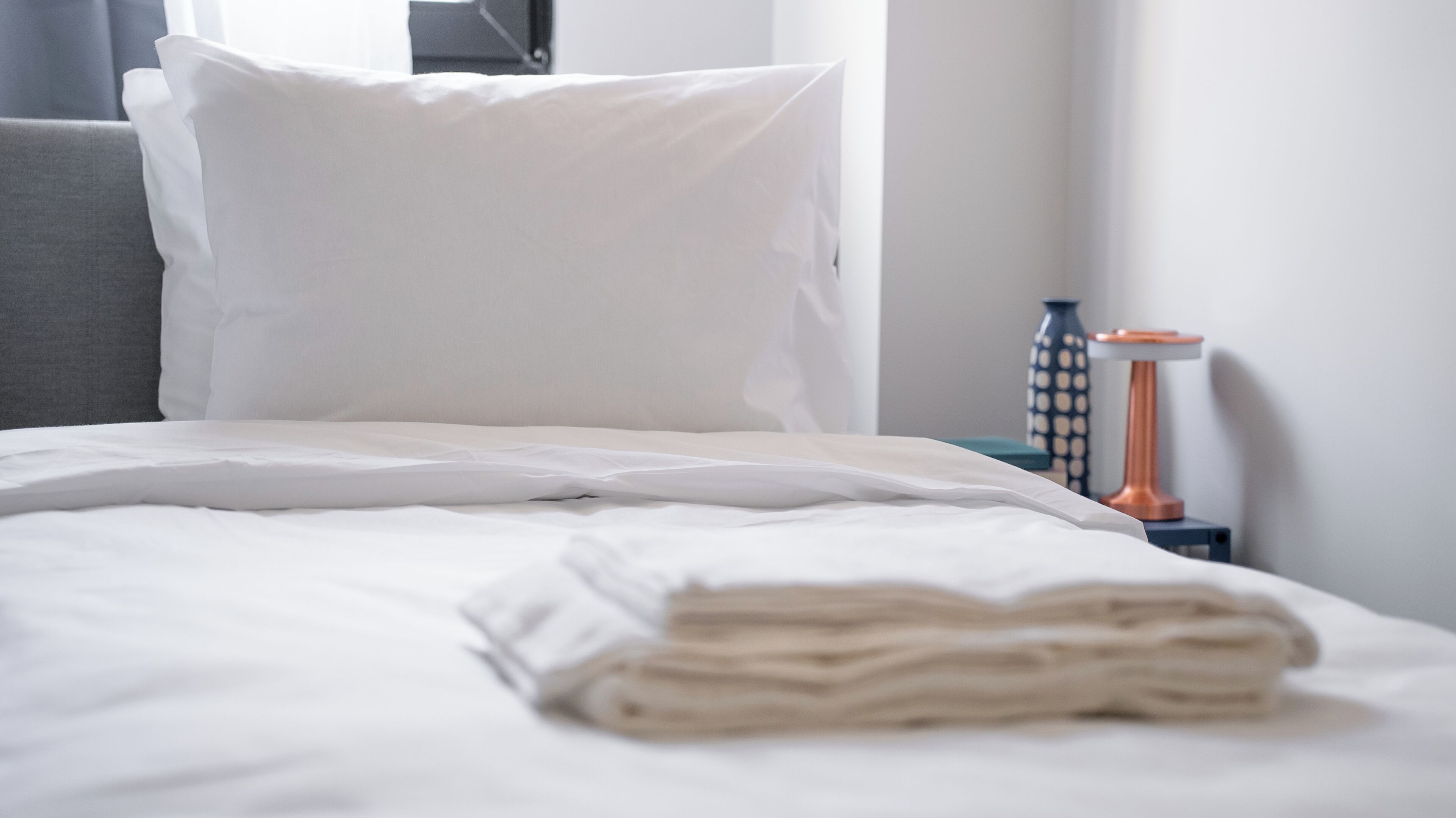 A neatly made bed with white linens, a grey headboard, and a bedside table with a vase and a lamp.