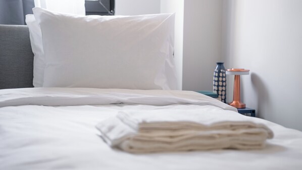 A neatly made bed with white linens, a grey headboard, and a bedside table with a vase and a lamp.